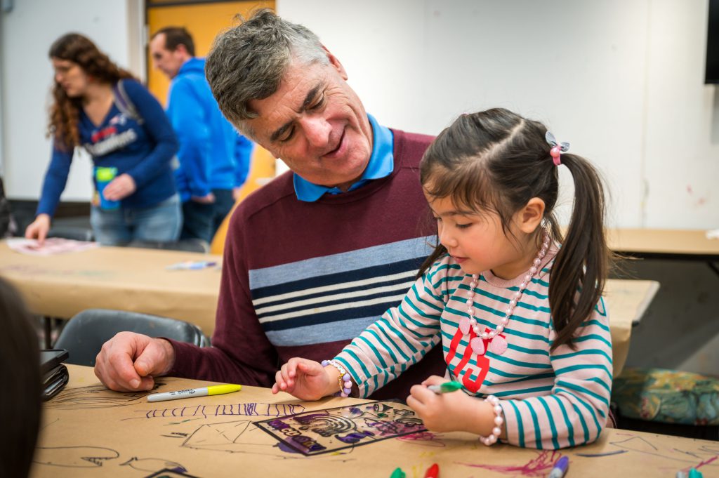 A young girl sits beside her caregiver, coloring an image of a menora