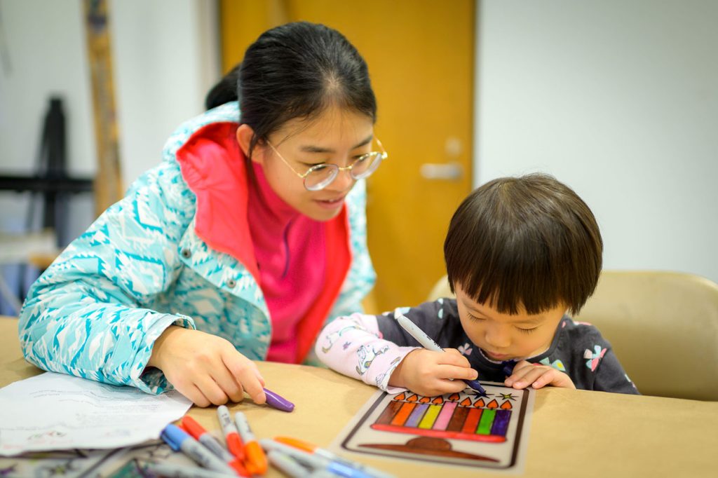 A young boy sitting at a desk coloring a picture of a menorah and a young girl stands watching him