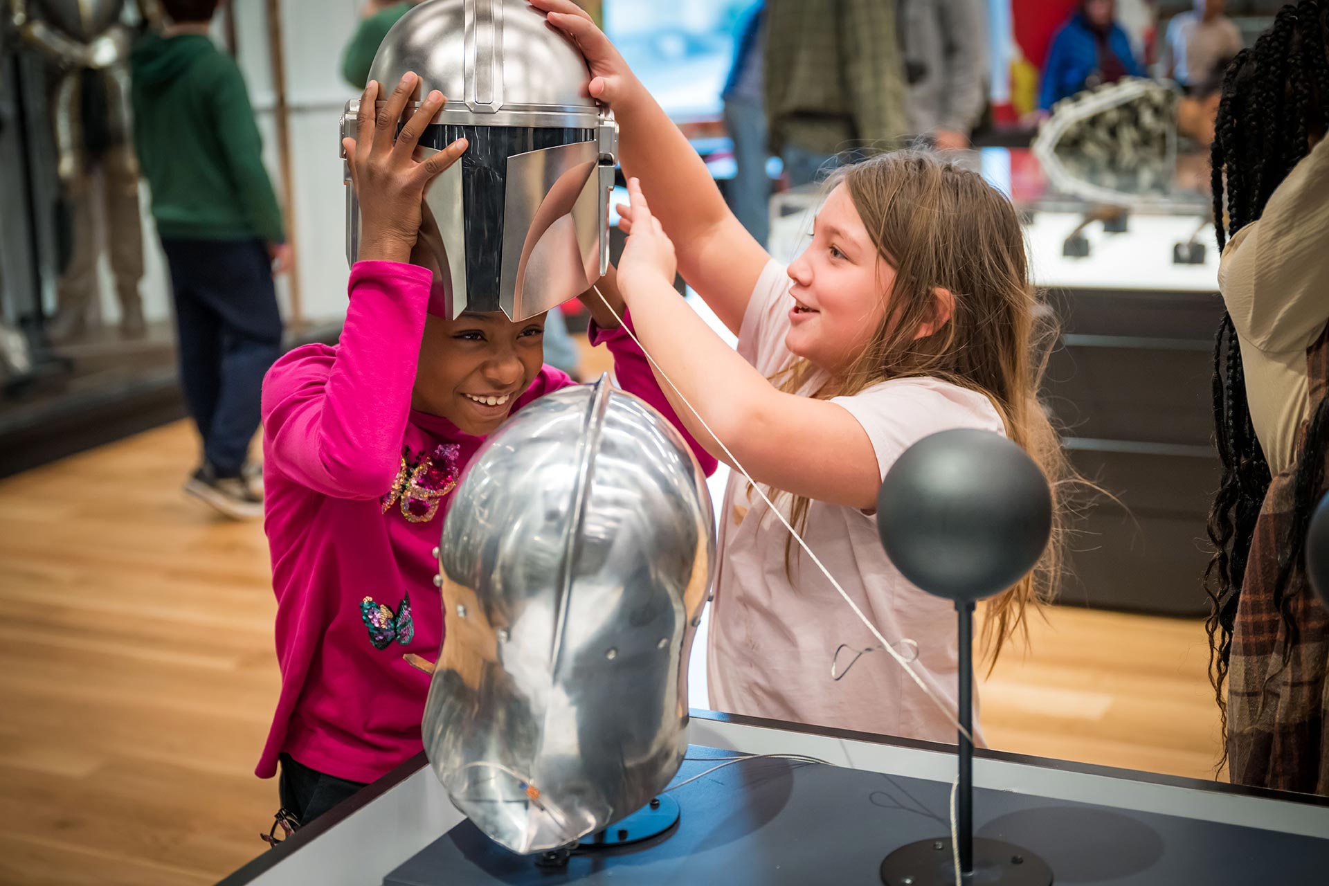 A young visitor tries on a helmet with the help of another