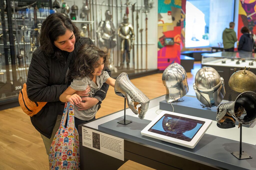 A young visitor is lifted up to reach inside a gauntlet in the Arms and Armor Galleries